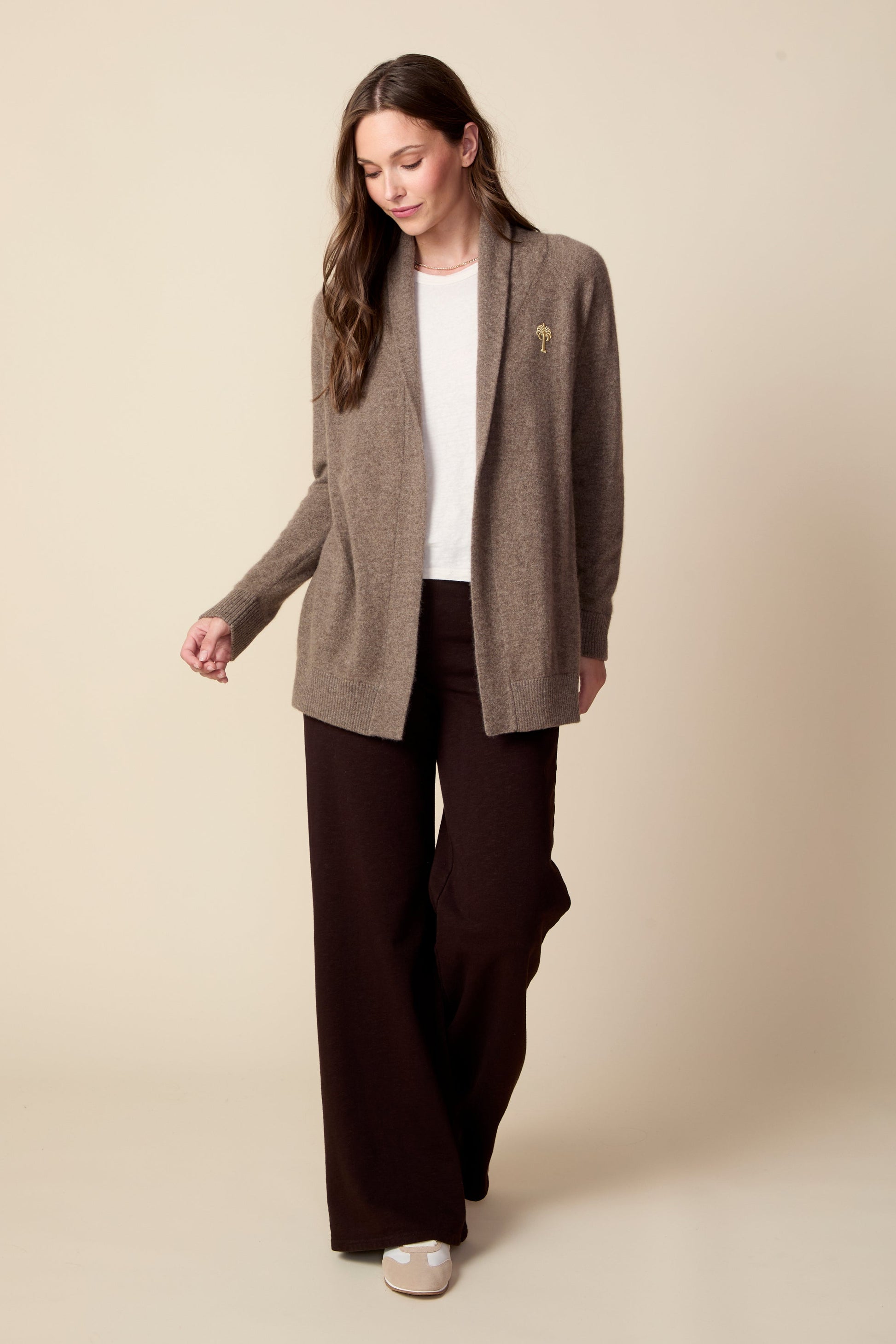 Woman wearing a brown cardigan over a white shirt and dark pants on a beige background
